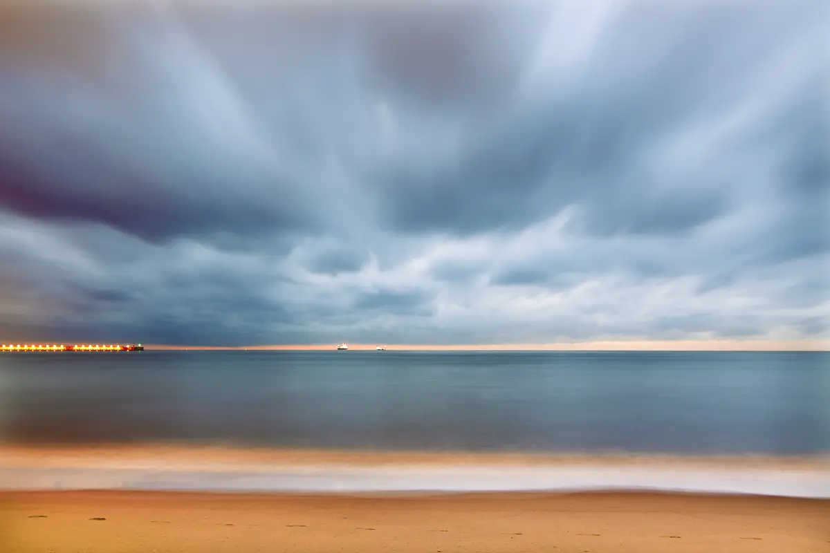 A lit marine terminal jetty and tankers offshore at dusk under storm clouds