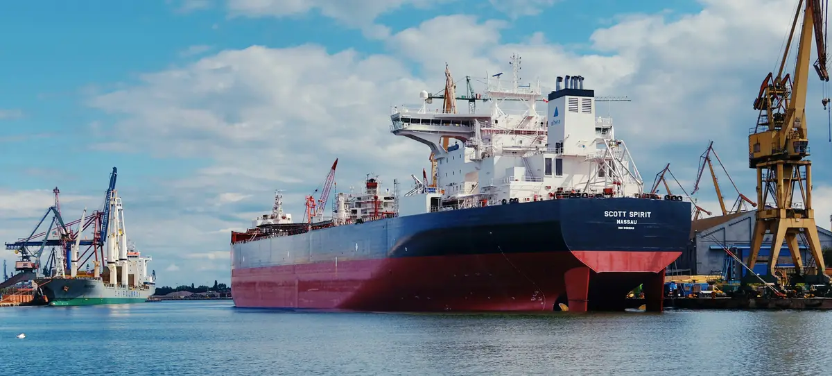 A tanker docked in port among cargo cranes under a partly cloudy sky